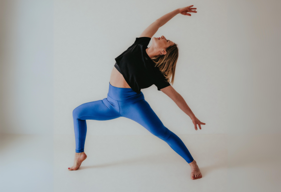 Eine Person in schwarzem Hemd und blauen Leggings führt eine Yoga-Pose aus und streckt sich seitwärts in einem hellen Raum.
